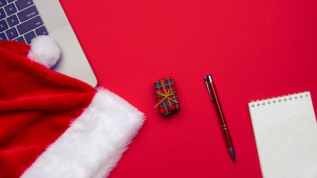 Christmas and Business Items on a red background, a cropped laptop, a Santa Claus cap, a pen, a notepad. Top view.