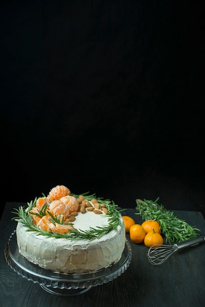 Christmas cake with tangerines, almonds and rosemary on a stand. Christmas cake with whipped cream. Dark background. Space for text.