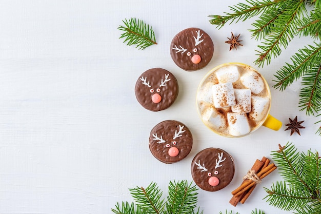 Christmas coffee with marshmallows and chocolate biscuits with deer faces on white background decorated with fir twigs.