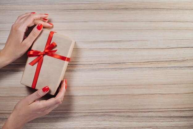 Christmas composition. Top view, flat lay. Female hands holding gift box wrapped in craft paper and decorated with red satin ribbon on wooden table