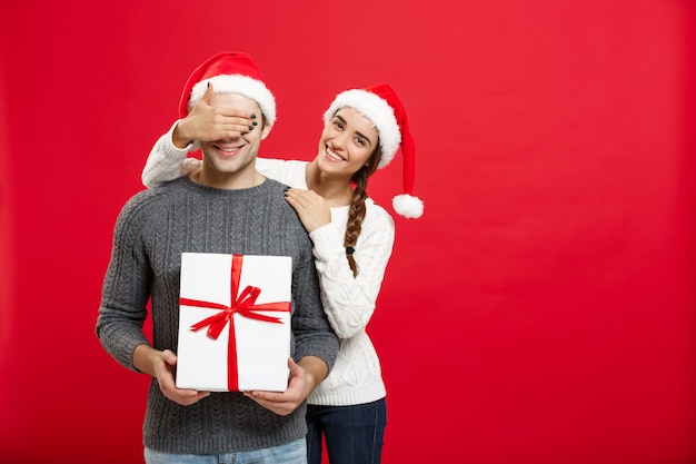 Christmas Concept - Young woman covering man's eyes with hand and giving surprise big gift. Isolated on Red wall.