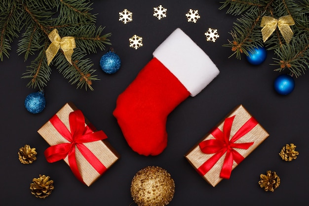 Christmas decoration. Santa's boot, gift boxes, fir tree branches with cones and christmas toys on black background. Top view. Christmas greeting card concept.