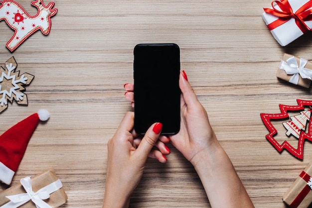 christmas decorations, pine tree toys and gifts on wooden table. female hands holding mobile phone with blank black screen. Top view.