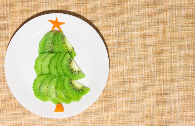 Christmas edible tree made from kiwi and carrot on plate on the table. Dessert idea for kids. New Year food art concept. Top view, copy space. High quality photo