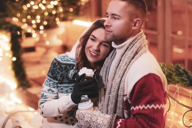 On the Christmas Eve charming couple of two romantic people are enjoying hot drinks while sitting on the bench.