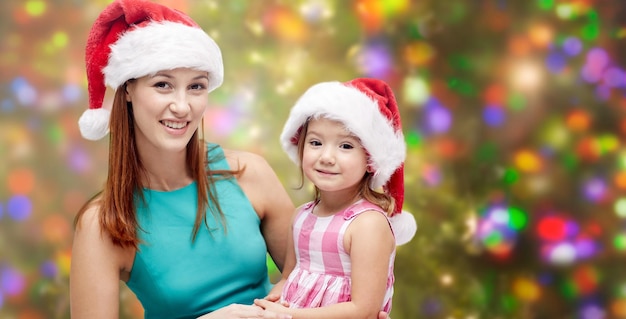 christmas, family, childhood and people concept - happy mother and little girl in santa hats over holidays lights background