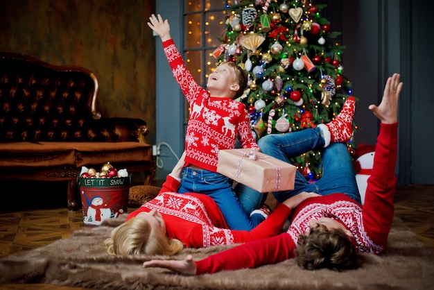 Christmas family portrait in red traditional sweaters in home holiday living room, parents and child with gift box.