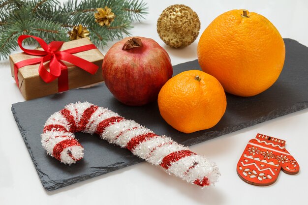 Christmas fir tree branch with cones, a gift box, an orange, a tangerine and a pomegranate on the stone board and the white background