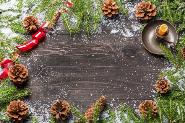 Christmas fir trees in snow with cones, red ribbon and candle in candlestick on a dark wooden board. Copy space for your text.