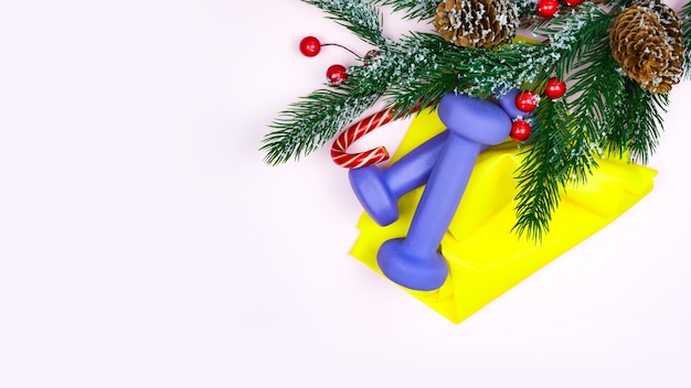 Christmas fitness. Healthy and active lifestyles  concept. Purple dumbbells, yellow rubber band, candy and fir-tree on pink