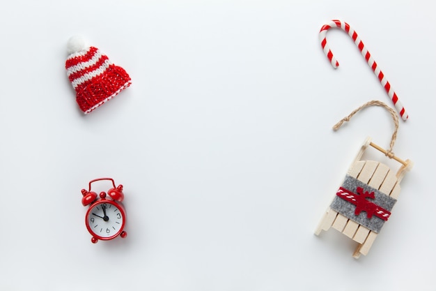 Christmas flat lay, winter beanie hat, candy cane, small red analog clock, sled on white background