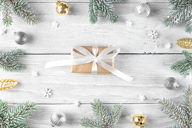 Christmas gift box in frame made of snowy fir tree, silver and golden decorations on white wooden background