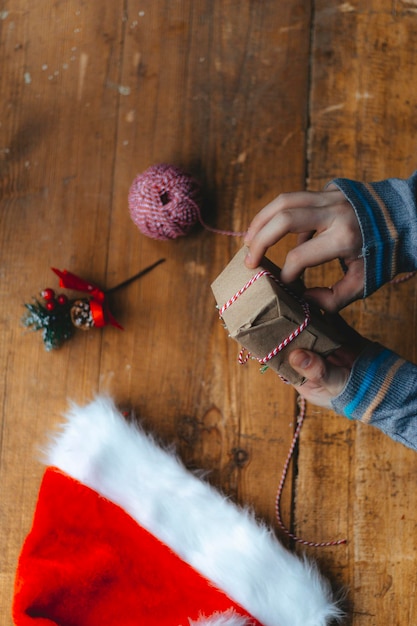 Christmas gift packaging. Hands wrap a small gift box in craft paper and a red-white string on the rustic wooden table with a Santa hat. Atmospheric image, Scandinavian nordic style.