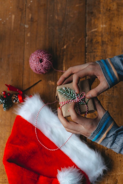 Christmas gift packaging. Hands wrap a small gift box in craft paper and a red-white string on the rustic wooden table with a Santa hat. Atmospheric image, Scandinavian nordic style