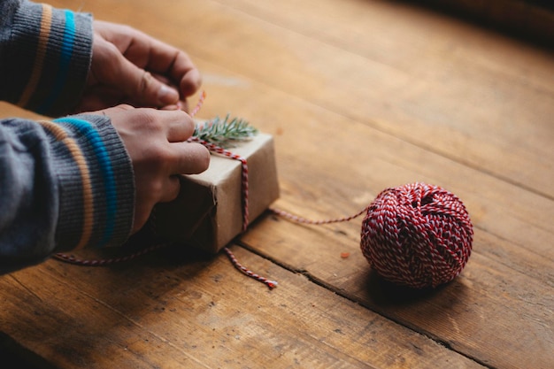 Christmas gift packaging. Men's hands in a blue sweater tie a bow on a small gift box on a wooden table. Selective focus