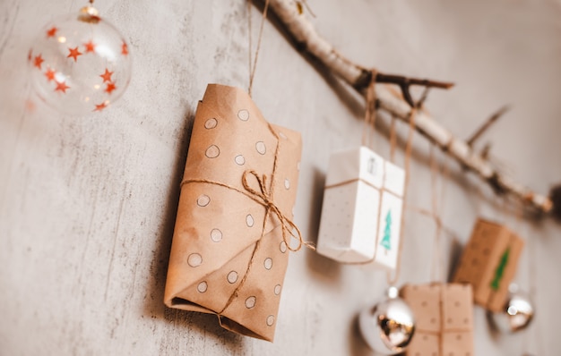 Photo christmas gifts packed with kraft paper and hand made decorations hang on a rope tied to a stick against a gray concrete wall.