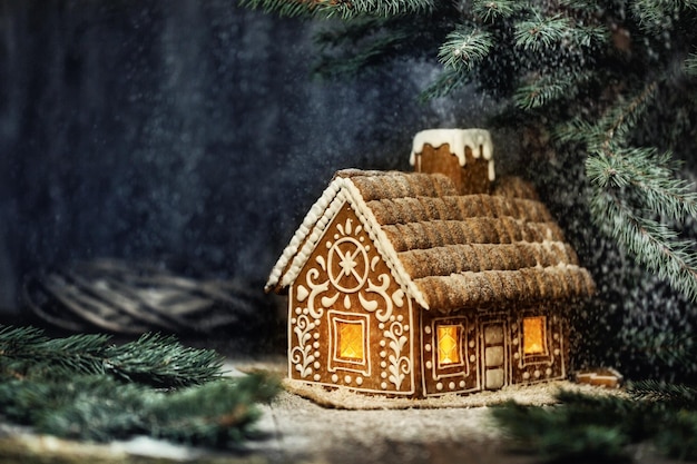 Christmas Gingerbread House with Window Lights in Winter Snowy Forestat Night. Creative Food Decoration Design for Xmas Holiday over Dark Background with Copy Space