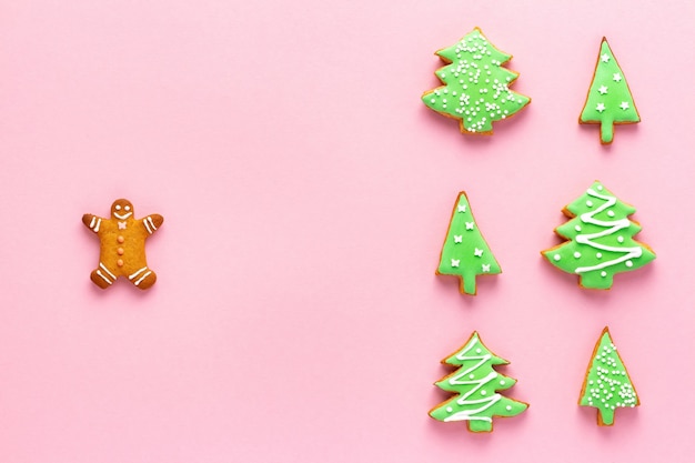 Christmas gingerbread man and a group of fir tree shaped cookies decorated with holiday icing on pink background, flat layout.