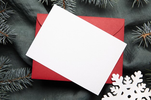 Christmas greeting card mockup with fir tree branches red envelope and white snowflake on green background top view