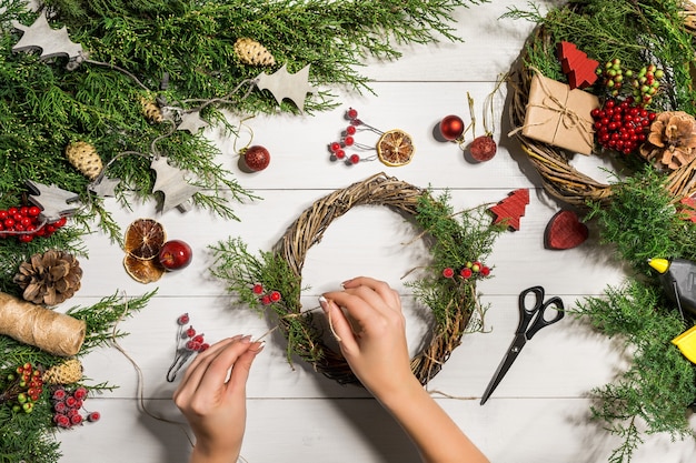 Christmas handmade diy background. Making craft xmas wreath and ornaments. Woman's leisure, tools and trinkets for holiday decorations. Top view of white wooden table with female hands.