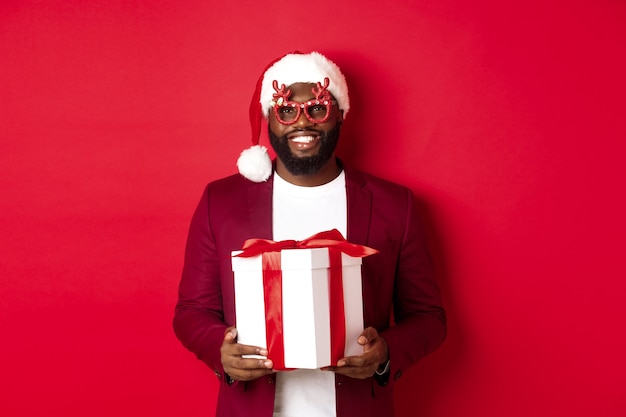 Christmas. Handsome african american man in party glasses and santa hat holding new year gift, bring present in box and smiling, standing over red background