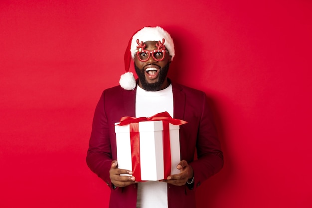 Christmas. Handsome african american man in party glasses and santa hat holding new year gift, looking surprised, receive present and smiling, standing over red background