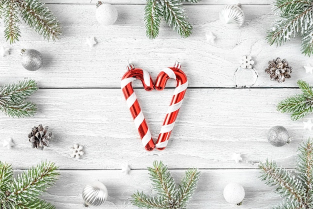 Christmas or Happy New Year composition. Candy in heart shape in frame made of fir tree branches, holiday decorations on white wooden background. Flat lay. Top view