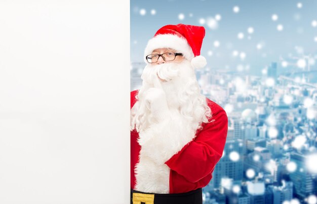 christmas, holidays, advertisement and people concept - man in costume of santa claus with white blank billboard making hust gesture over snowy city background