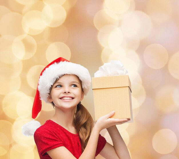 christmas, holidays, childhood and people concept - smiling girl in santa helper hat with gift box over beige lights background