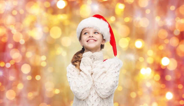 christmas, holidays and children concept - dreaming girl in santa helper hat over lights background