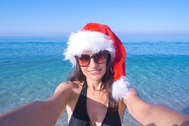 Christmas and holidays concept Beautiful woman in Santa's hat takes a selfie on the seashore