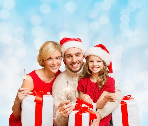 christmas, holidays, family and people concept - happy mother, father and little girl in santa helper hats with gift boxes and sparklers over blue lights background