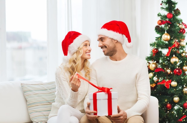 christmas, holidays and people concept - happy couple in santa hats with gift box sitting on sofa at home