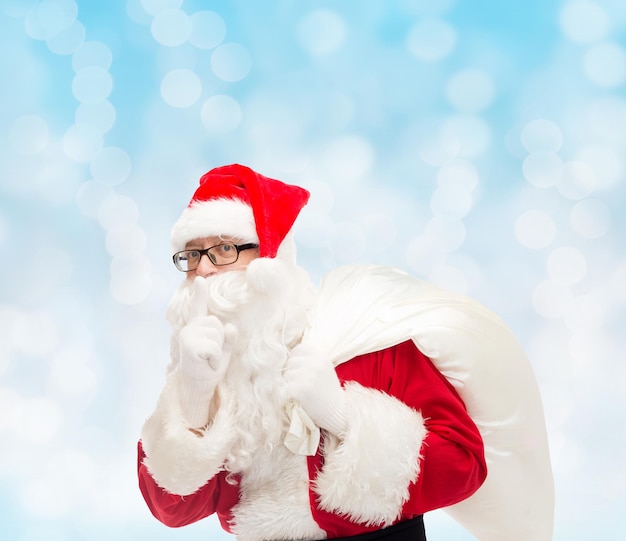 christmas, holidays and people concept - man in costume of santa claus with bag making hush gesture over blue lights background