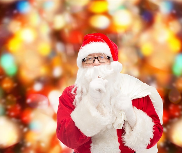 christmas, holidays and people concept - man in costume of santa claus with bag making hush gesture over red lights background