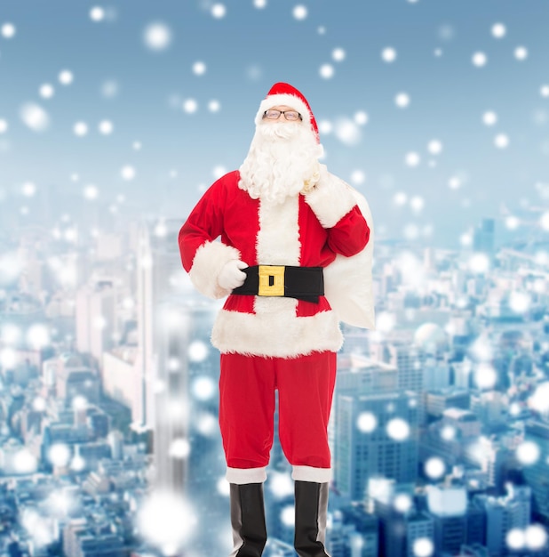 christmas, holidays and people concept - man in costume of santa claus with bag over snowy city background