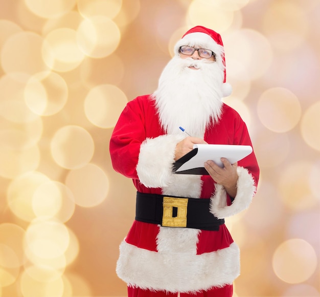 christmas, holidays and people concept - man in costume of santa claus with notepad and pen over beige lights background