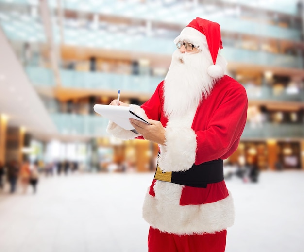 christmas, holidays and people concept - man in costume of santa claus with notepad and pen over shopping center background