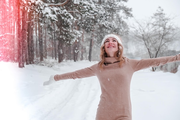 Christmas, holidays and season concept. Young happy woman blowing snow in the winter forest nature. Warm clothing knitted gloves and hat. Winter forest landscape background