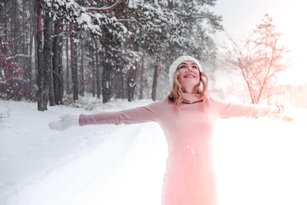 Christmas, holidays and season concept. Young happy woman blowing snow in the winter forest nature. Warm clothing knitted gloves and hat. Winter forest landscape background