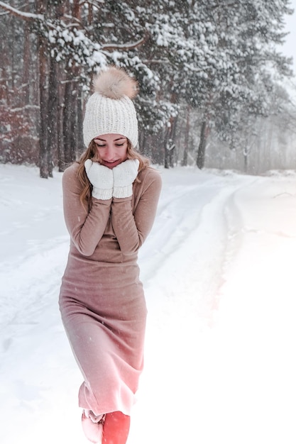 Christmas, holidays and season concept. Young happy woman blowing snow in the winter forest nature. Warm clothing knitted gloves and hat. Winter forest landscape background