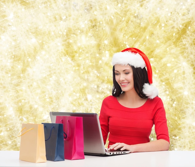 christmas, holidays, technology and people concept - smiling woman in santa helper hat with shopping bags and laptop computer over yellow lights background