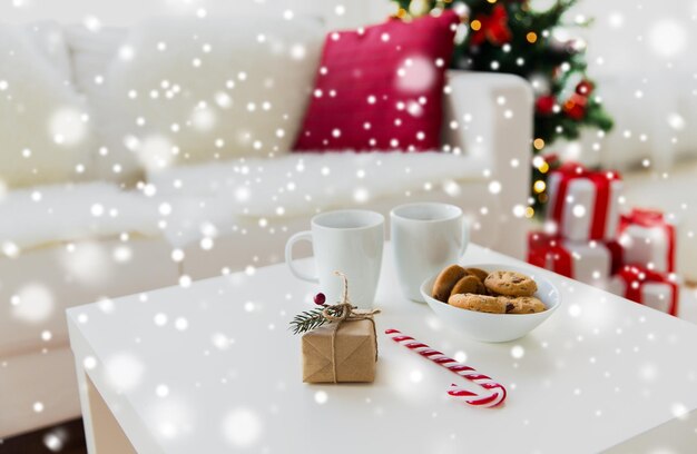 christmas, holidays and  winter concept - close up of gift, oat cookies, sugar cane candy and cups on table at home