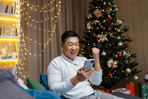 Christmas man near tree at home reading good news from phone got win and discount in online casino