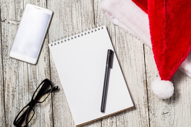 Christmas and New Year background with smartphone, red Santa hat, glasses, notepad and pen for your wishes