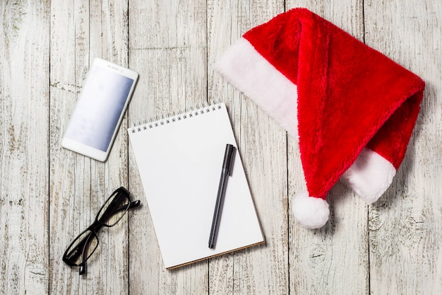 Christmas and New Year background with smartphone, red Santa hat, glasses, notepad and pen for your wishes