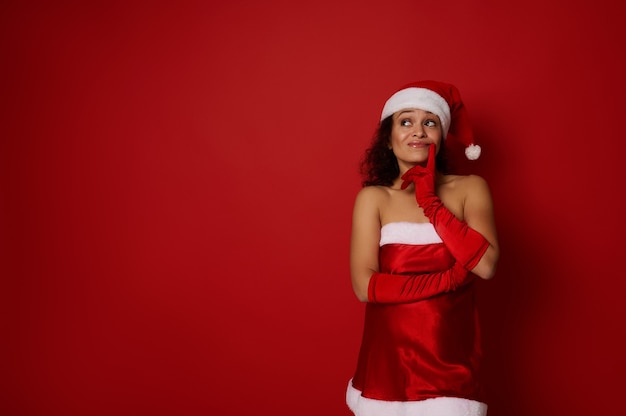 Christmas and New Year concept for ad. Pensive mysterious beautiful woman in Santa carnival costume holds her finger near her lips and looks thoughtfully up at a copy space for ad on red background.