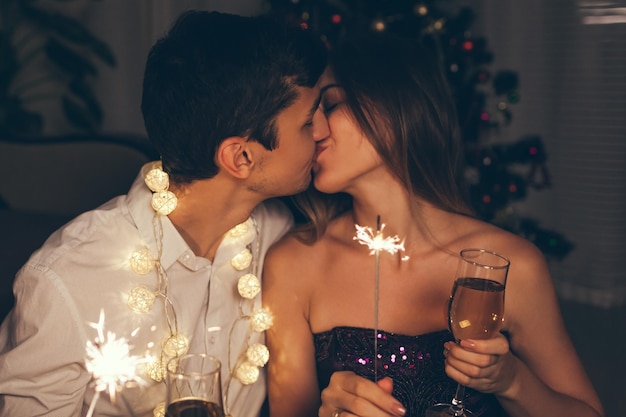 Christmas and New year masquerade party concept. Couple kissing, burning sparklers by illuminated Christmas tree with champagne at home. Celebrating holidays