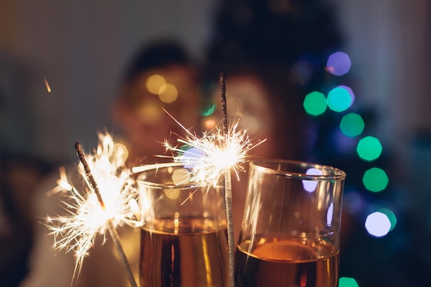 Christmas and New year masquerade party concept. Couple in love burning sparklers by illuminated Christmas tree with champagne at home.