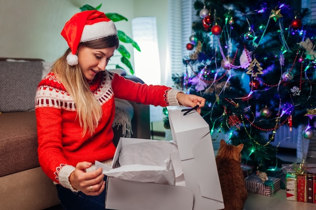 Christmas and New Year presents Woman in Santas hat opening gift box by Christmas tree at home